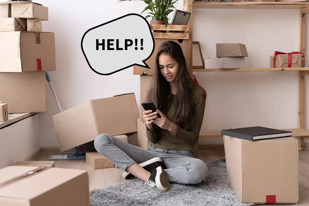 A student sits on a rug in a room filled with packed cardboard boxes, using her smartphone to book self storage for students. Beside her is a stack of boxes and a cleaning mop, illustrating the transition of moving out of university accommodation.